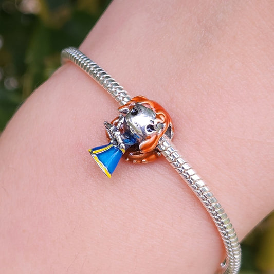 Ajoutez une touche de courage et de magie à votre collection avec ce charm inspiré de Merida, l’héroïne audacieuse du film Rebelle. Fabriqué en métal argenté et émaillé de nuances de bleu et de vert, ce charm capture l'esprit intrépide de Merida avec des détails représentant son arc et sa chevelure flamboyante. Orné de motifs celtiques et d’un subtil éclat qui rappelle les feux follets de la forêt enchantée, ce charm est parfait pour les amoureux des contes de fées modernes et des aventures épiques.