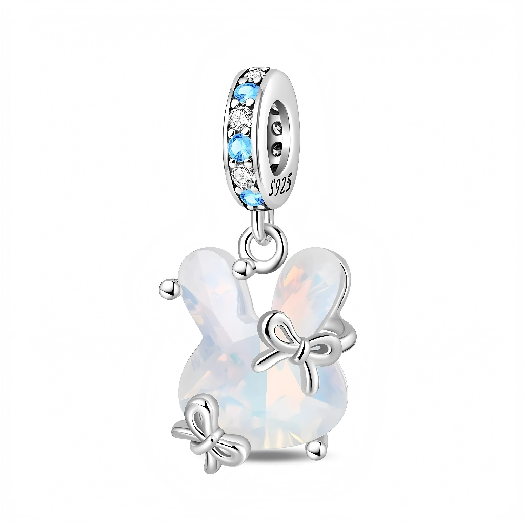 Ce charm pendant en argent 925 représente un petit lapin stylisé entièrement formé d’une pierre synthétique opalescente aux reflets laiteux et irisés, lui donnant un aspect doux et lumineux. Trois petits nœuds en argent viennent s’y poser, tandis que la bélière est soigneusement pavée d’oxydes de zirconium blancs et de cristaux bleu clair pour une touche fraîche et scintillante.