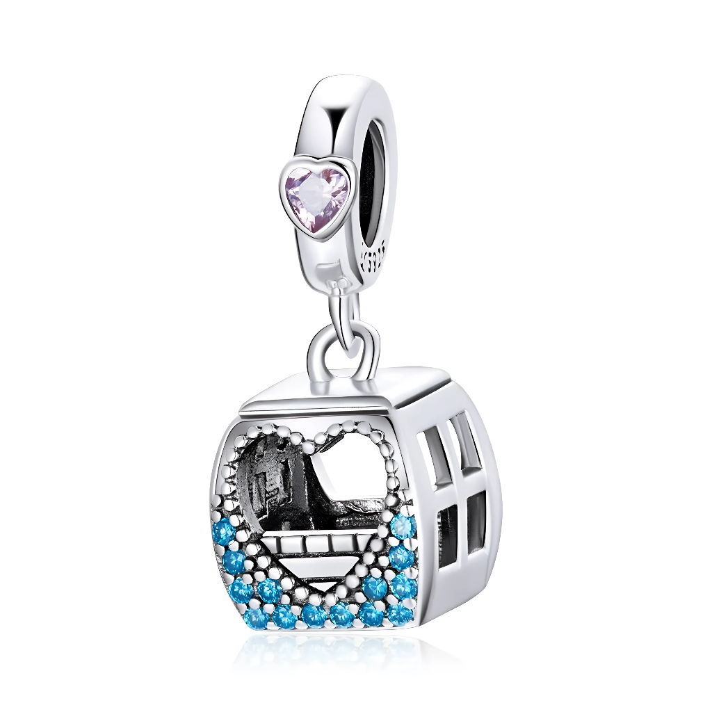 La bélière est décorée d’un petit cristal en forme de cœur rose clair, ajoutant une touche romantique au thème du voyage. La cabine carrée ajourée montre, à travers une ouverture en forme de cœur sertie de pierres bleues, une scène miniature de paysage urbain ou montagneux, comme si l’on regardait par la fenêtre du téléphérique. Les côtés sont découpés de fenêtres, ce qui renforce l’effet de petite nacelle suspendue.