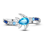 Cette bague en argent 925 met à l’honneur une adorable tortue de mer, sertie d’un cristal bleu en forme de goutte qui évoque immédiatement la transparence des lagons tropicaux. Les petits oxydes de zirconium dégradés du bleu nuit au bleu clair et au blanc se prolongent sur l’anneau, comme une traînée d’écume, pour un bijou lumineux qui rappelle la liberté de l’océan à chaque mouvement de la main.